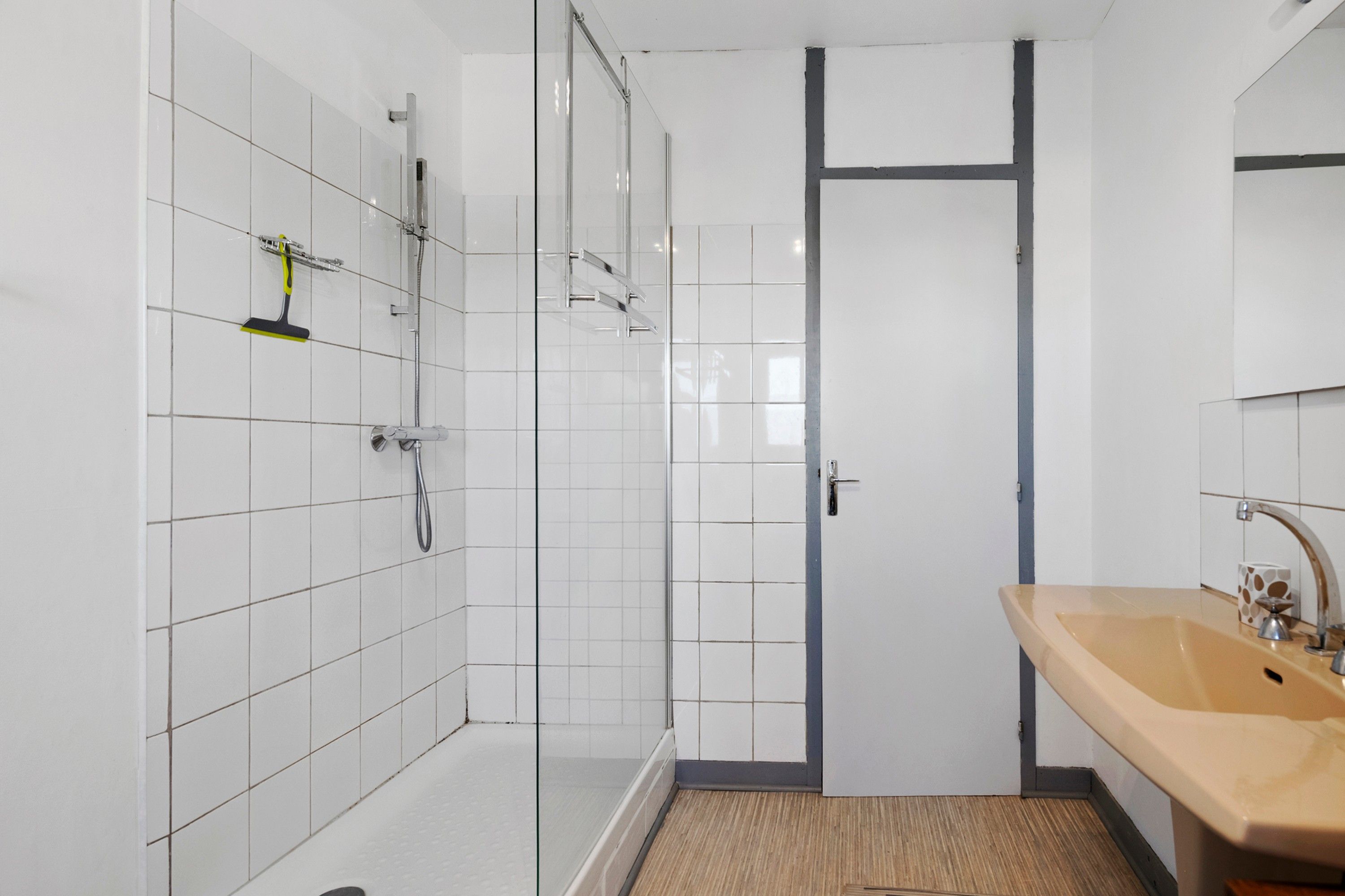 Ein Badezimmer mit weiß gefliesten Wänden, einer Dusche, einem Waschbecken und einer Tür.