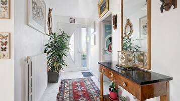Un couloir avec un radiateur, une plante en pot, une console en bois sur laquelle se trouve une horloge, et un tapis à motifs.