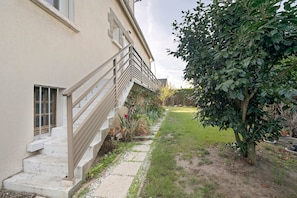 Un edificio con una escalera de piedra que lleva a una puerta, una barandilla de metal y un jardín con un árbol y césped.
