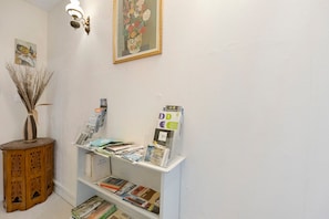 Un área de recepción con una mesita de madera, un florero con plantas secas, una lámpara de pared, un cuadro con motivos florales y un expositor con folletos.