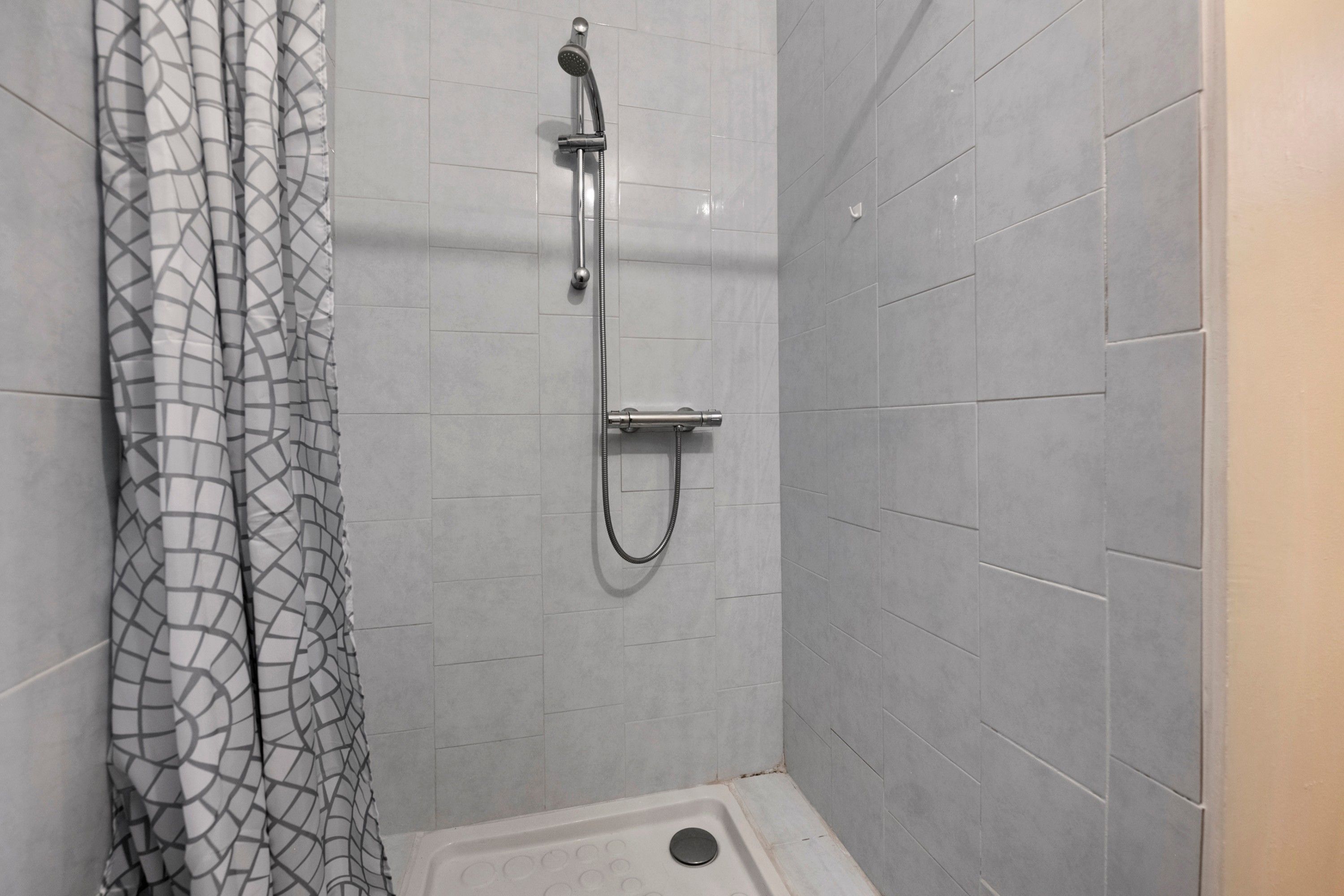 Eine Dusche mit Handbrause und Wandarmatur, ein Duschvorhang mit geometrischem Muster sowie ein gefliester Boden.