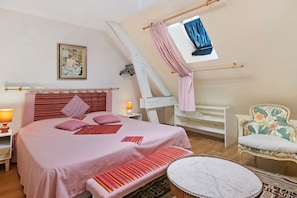 Un dormitorio con cama, mesitas de noche, un banco, una silla, una ventana con cortinas y un cuadro en la pared.
