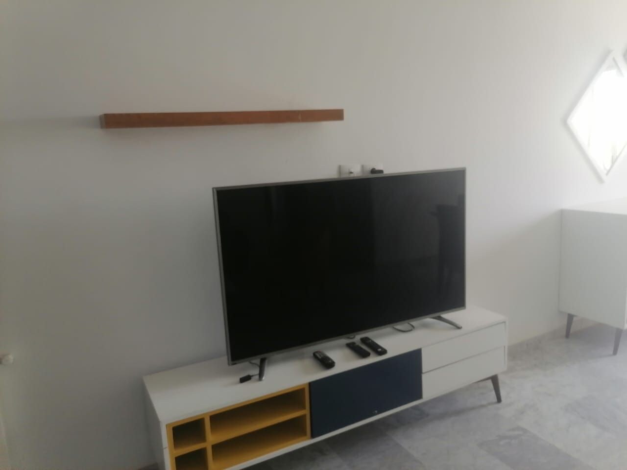 Un salon moderne avec un téléviseur à écran plat posé sur un meuble blanc, une étagère en bois au-dessus et un radiateur blanc fixé au mur.