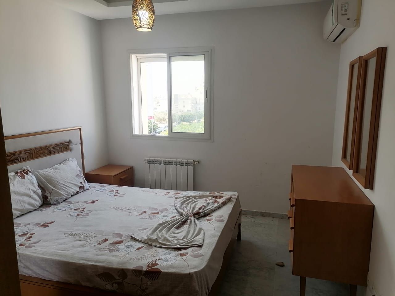 Een slaapkamer met een bed, nachtkastje, radiator, raam en een kast.
