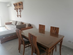 A dining area with a glass-top table and wooden chairs, a sofa, and a wall-mounted decorative piece. - Fully Furnished 1-bedroom Apartment in Khezama Prime Location (Sousse)
