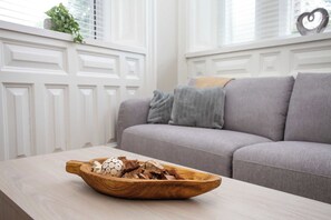 Eine Holzschüssel auf einem Tisch mit verschiedenen Gegenständen, ein graues Sofa und ein Fenster mit Jalousien in einem Raum.