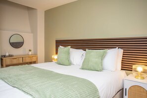 Un lit bien fait, recouvert d’un plaid vert, une table de chevet avec une lampe et un miroir fixé au mur.