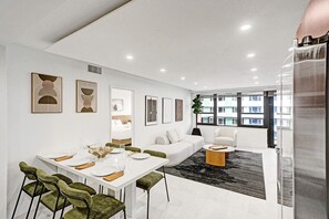 A modern living room with a white sofa, a dining table set for four, and a large window offering a view of buildings. - Spacious 3BR Retreat Steps from the Beach with Resort Amenities 1508 (Miami Beach)