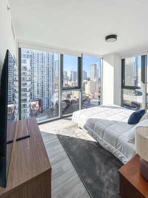 Une chambre moderne avec un grand lit, un bureau en bois et une vue sur le paysage urbain grâce à de grandes fen êtres du sol au plafond.