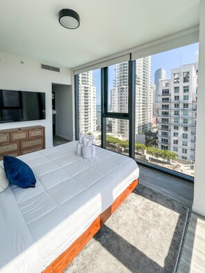 Ein modernes Schlafzimmer mit einem großen Bett, einem Fernseher und Blick auf hohe Gebäude.