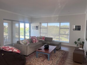 A spacious living room with a grey sofa, a coffee table, and a large window offering a view of the outdoors. - Gorgeous 4-bedroom house in charming Onrusrivier perfect for families. (Onrusrivier)