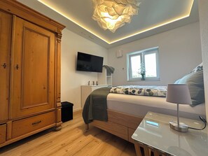 A bedroom with a bed, bedside table, lamp, TV, wardrobe, and a window with a view. - Comfortable Vacation Rental with Infrared Sauna & Wi-Fi Near Lake Sorpe | Haus am Krähenbach-Krähenblick (Sundern (Sauerland))