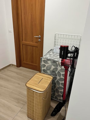 Ein Zimmer mit Holztür, einem gemusterten Bett, einem roten Föhn und einem Korbwägelchen auf dem Boden.