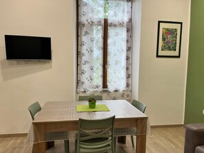 Ein Essbereich mit einem Tisch, Stühlen, einem an der Wand montierten Fernseher und einem Fenster mit blumigen Vorhängen.