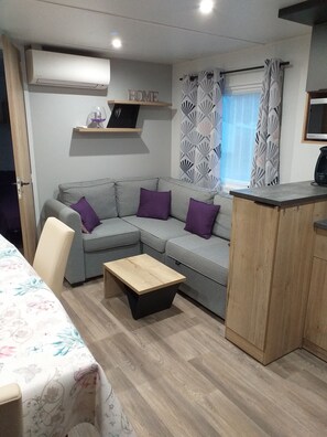 Ein modernes Wohnzimmer mit einem grauen Sofa, violetten Kissen, einem Holztisch, einer kleinen Holzkommode und einer Wand-Klimaanlage.