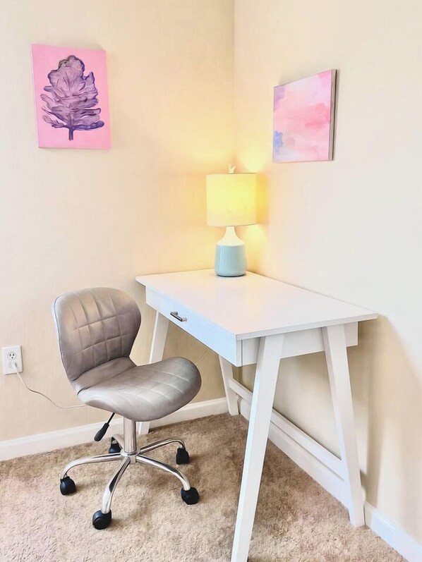 A corner room with a white desk, a cushioned office chair, a lamp, and two wall-mounted artworks. - Modern Charlotte Apt - Pool, Gym, Game Room, & BBQ (Charlotte)