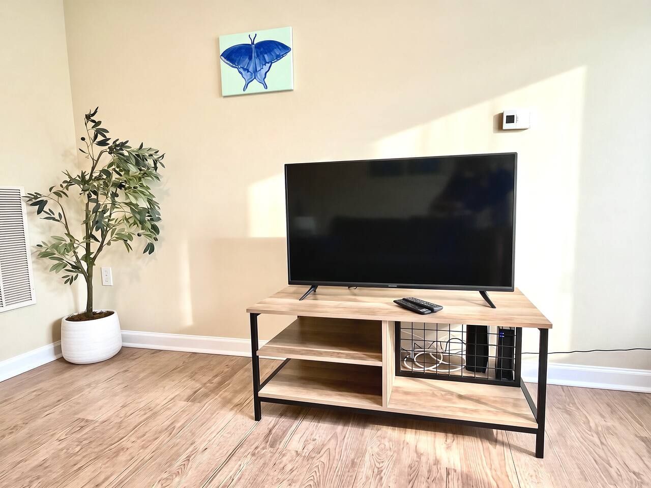Un televisor de pantalla plana sobre un mueble de televisión de madera, con un control remoto, una planta en maceta y una decoración de mariposa en la pared en una habitación con piso de madera.