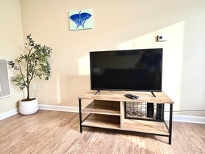 A flat-screen TV on a wooden TV stand with a remote, a potted plant, and a butterfly wall art in a room with wooden flooring. - Modern Charlotte Apt - Pool, Gym, Game Room, & BBQ (Charlotte)