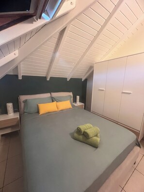 Ein Schlafzimmer mit schrägen Wänden, einem Bett mit grün-gelber Bettwäsche, einem Nachttisch und einem Kleiderschrank.