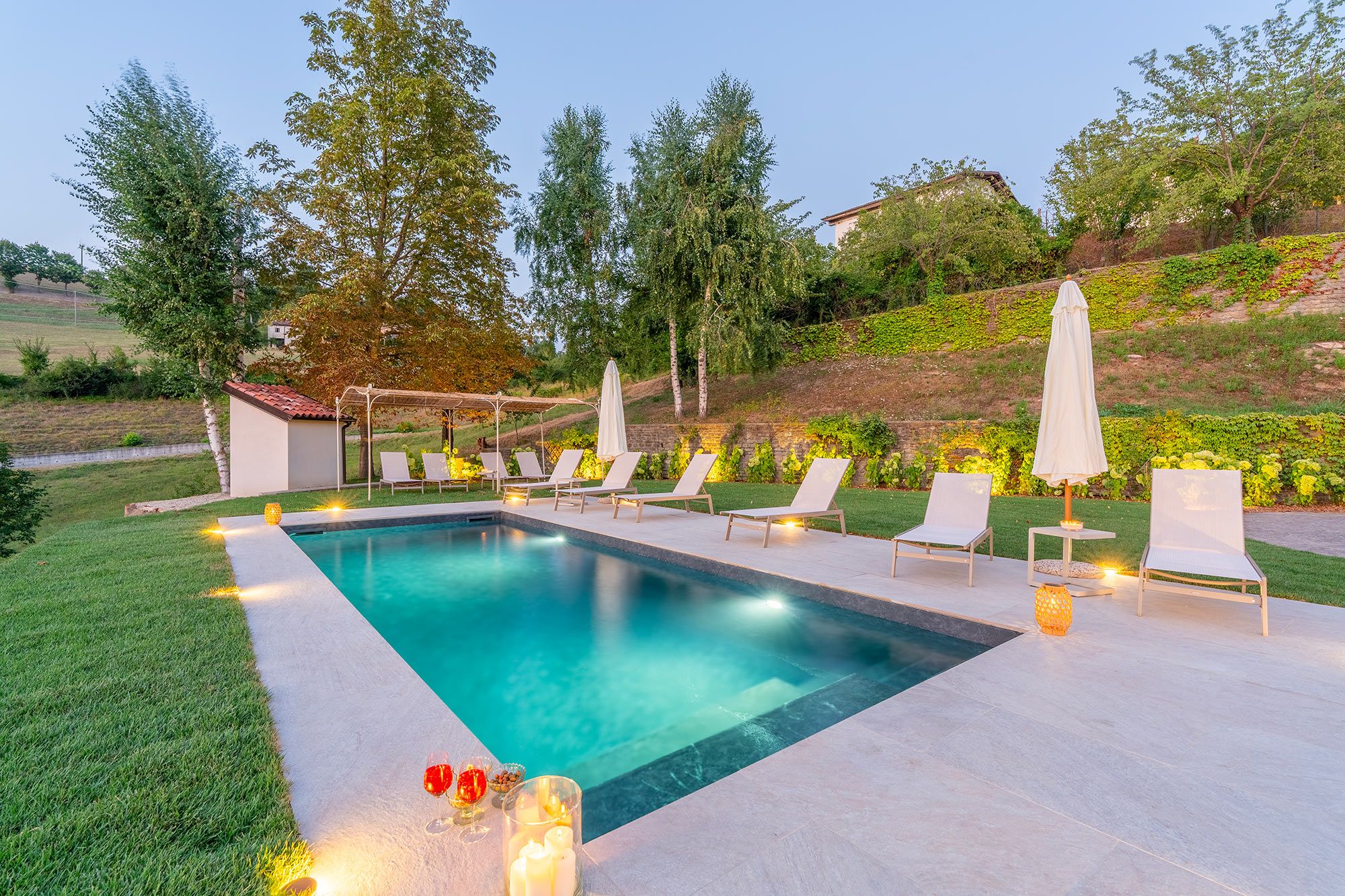 Une piscine entourée de verdure et bordée d’une colline, avec des chaises longues et des parasols.