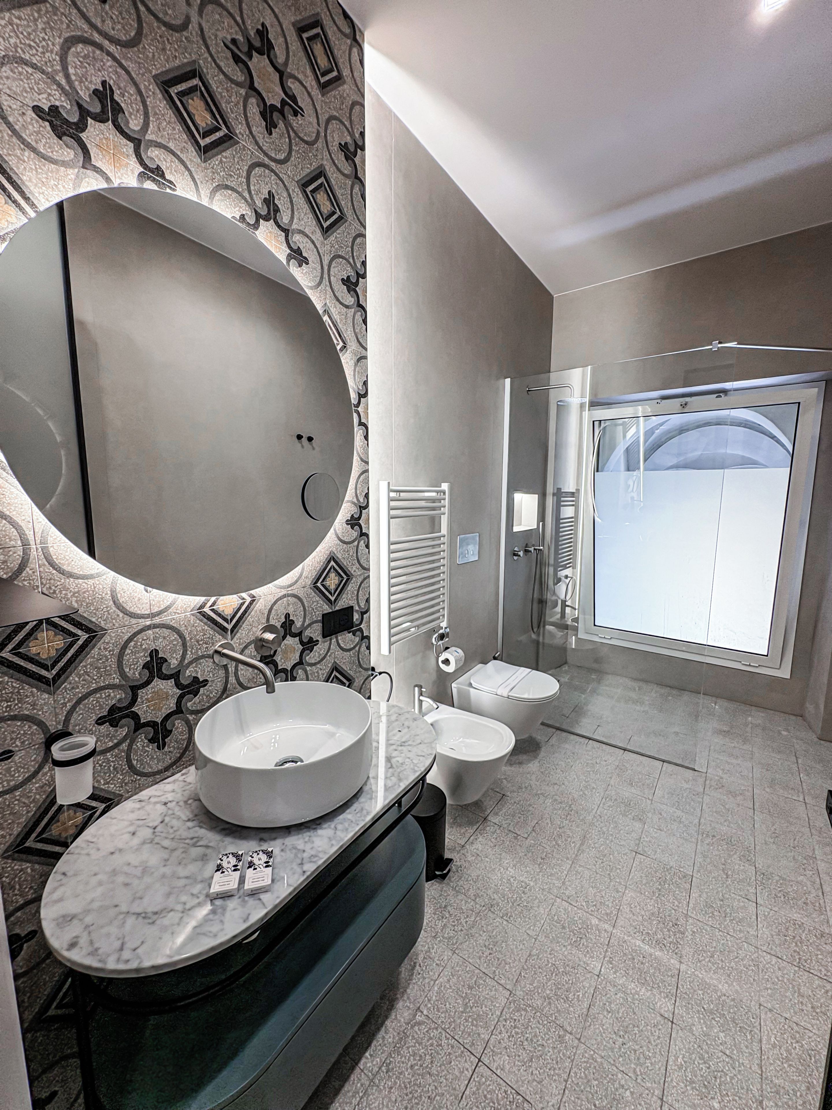 Un bagno moderno con un ampio specchio ovale, due lavandini, un water e una cabina doccia.