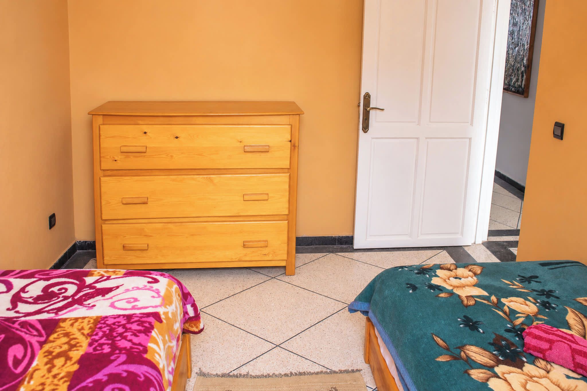 Une pièce avec une commode en bois, une porte blanche et deux lits avec une literie aux couleurs vives.