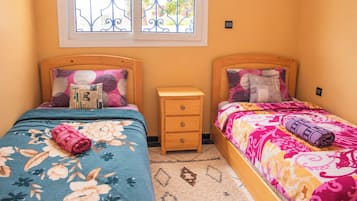 Une chambre avec deux lits, une petite table de chevet en bois et une fenêtre ornée de barreaux en fer.