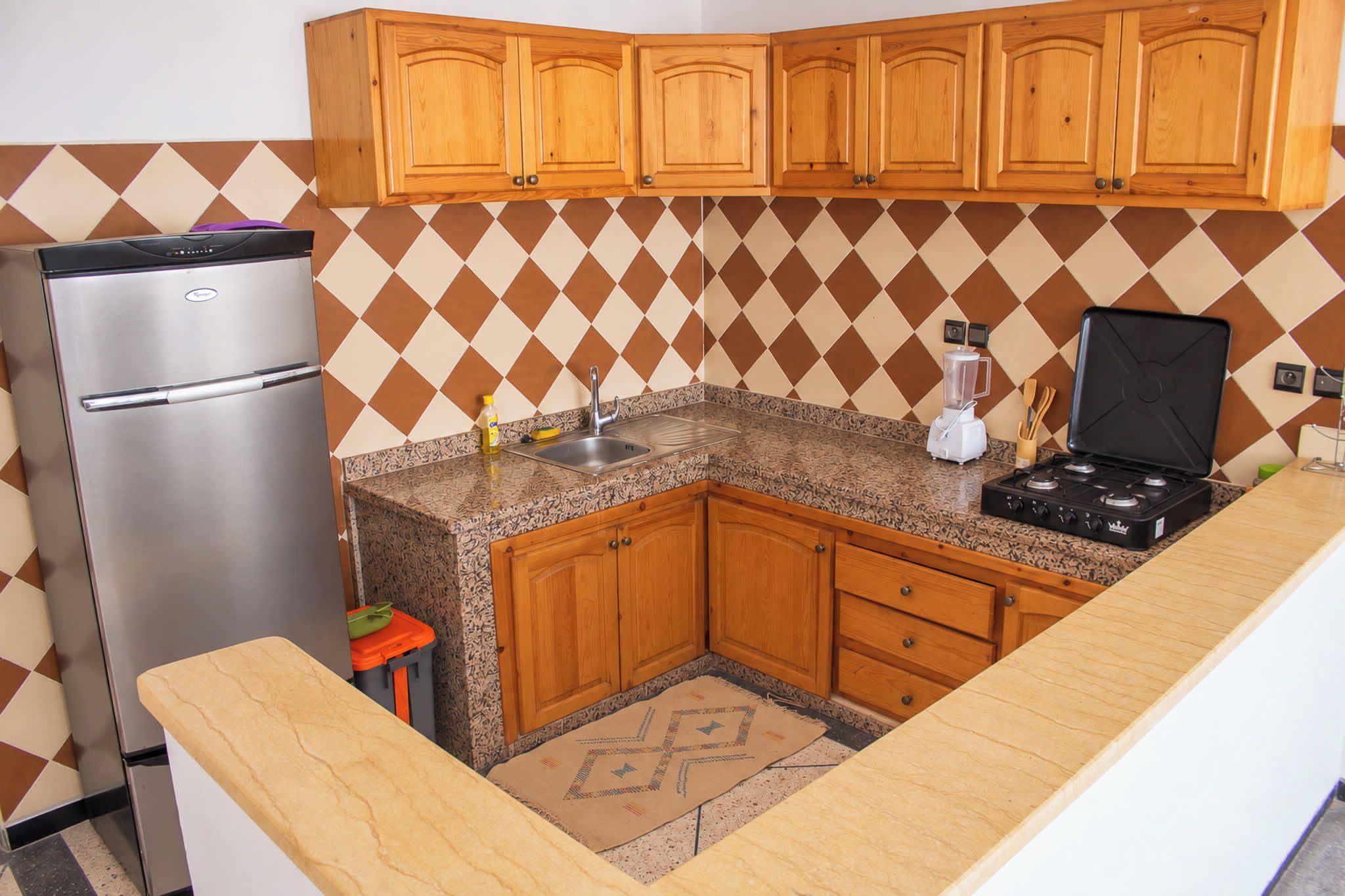 A compact kitchen with wooden cabinets, a stainless steel refrigerator, a countertop with a blender, and a patterned rug.