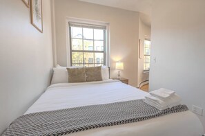 A neatly made bed with pillows, a bedside lamp, and a window with blinds. - Expansive South End Studio (Boston)