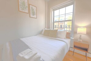 A bedroom with a bed, a nightstand, a window, and two wall-mounted plant illustrations. - Expansive South End Studio (Boston)
