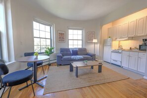 A living room with a sofa, coffee table, and chairs, a dining area with a table and chairs, and a kitchen with cabinets and appliances. - Expansive South End Studio (Boston)