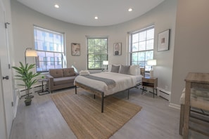 A bedroom with a bed, a sofa, a nightstand, a lamp, and a potted plant. - Spacious, Light-Filled Studio in the South End (Boston)
