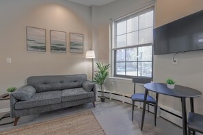 A living room with a grey sofa, a small table, a floor lamp, and a window with blinds. - Bold Studio Steps from Back Bay (Boston)