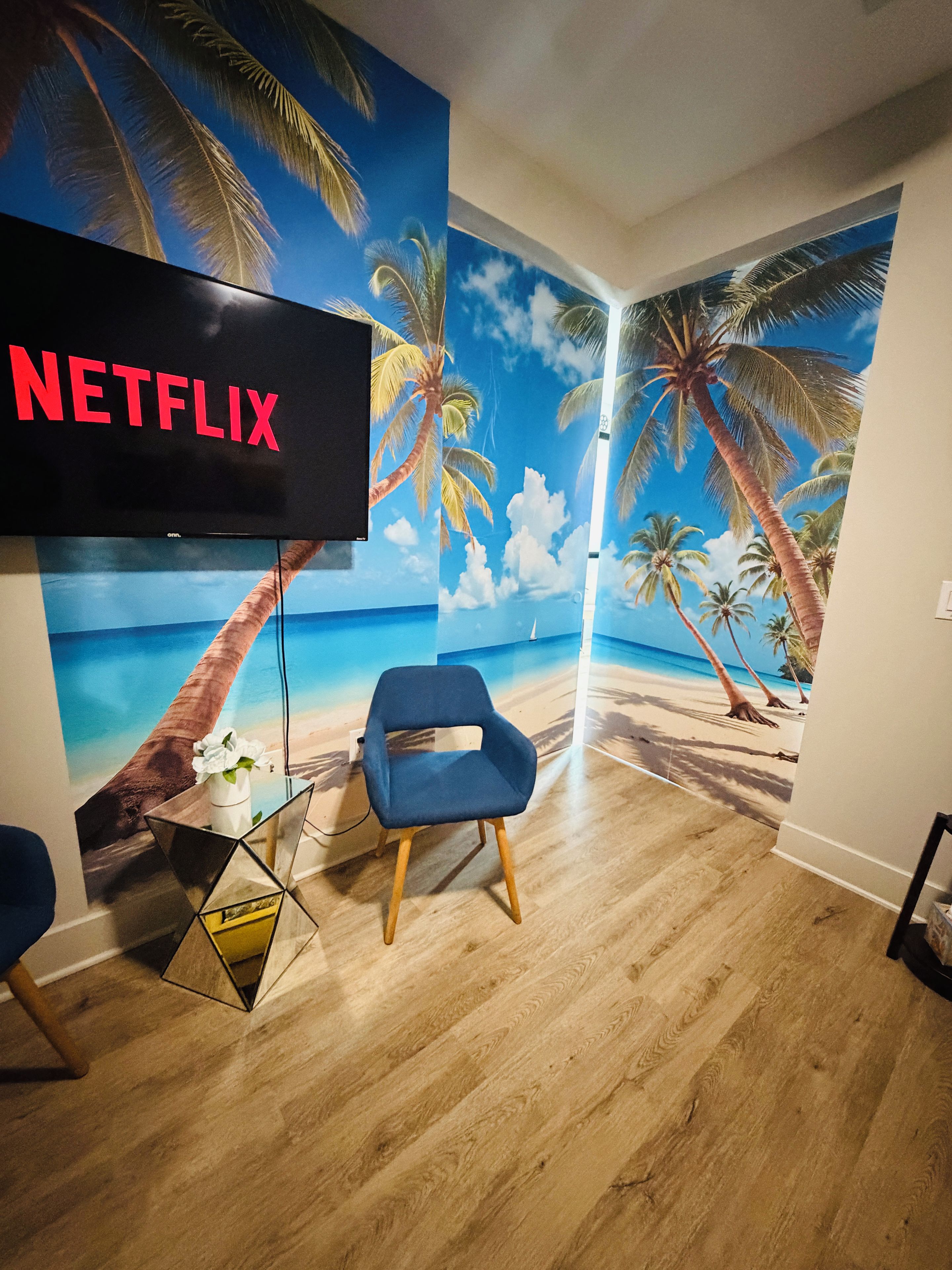 A room with a wall mural of a beach scene, a flat-screen TV mounted on the wall, a blue chair, and a small table with a vase.