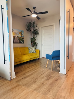 A room with a yellow sofa, a blue chair, a ceiling fan, a door, and a large plant. - Deep Ellum Theatre Suite – Private Cinema, Skyline & City Vibes (Dallas)