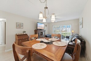 A dining area with a wooden table, chairs, a television, and a view of the outdoors through large windows. - Great NSB Location on Intercoastal (New Smyrna Beach)