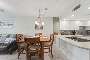 A modern kitchen with white cabinets, a central island with a stove, and a dining area with wooden chairs and a chandelier. - Great NSB Location on Intercoastal (New Smyrna Beach)