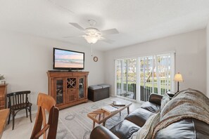A living room with a television, a ceiling fan, a sofa, a coffee table, and a view through a sliding glass door. - Great NSB Location on Intercoastal (New Smyrna Beach)