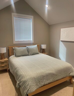 A bedroom with a sloped ceiling, a bed with a quilted bedspread, two bedside lamps, a window with blinds, and a wall-mounted artwork. - Cozy Okanagan escape between Big White & SilverStar - ski, relax & unwind! (Kelowna)