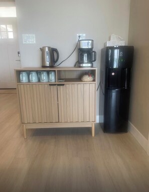 Eine Küchenzeile mit einem Holzkasten, einer Kaffeemaschine, einem Wasserkocher, einem Wasserspender und einem Kühlschrank.