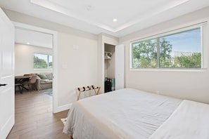 Un dormitorio con una cama grande, una ventana con persianas, una puerta que da a una sala de estar y un armario con ropa colgada.