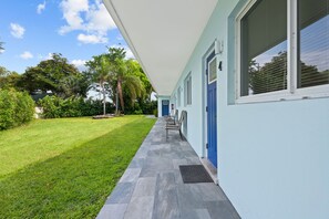 A building with a covered porch, a blue door, and a small garden area with grass and trees. - NEW LISTING: Charming 2-Bedroom Deerfield Beach Apartment Near Beach & Shops (Deerfield Beach)