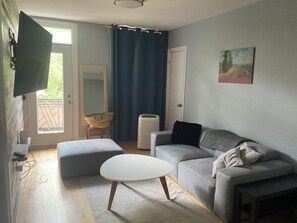 Ein Wohnzimmer mit grauem Sofa, weißem Hocker, einem flachbildfernseher an der Wand und einem Fenster mit blauen Vorhängen.