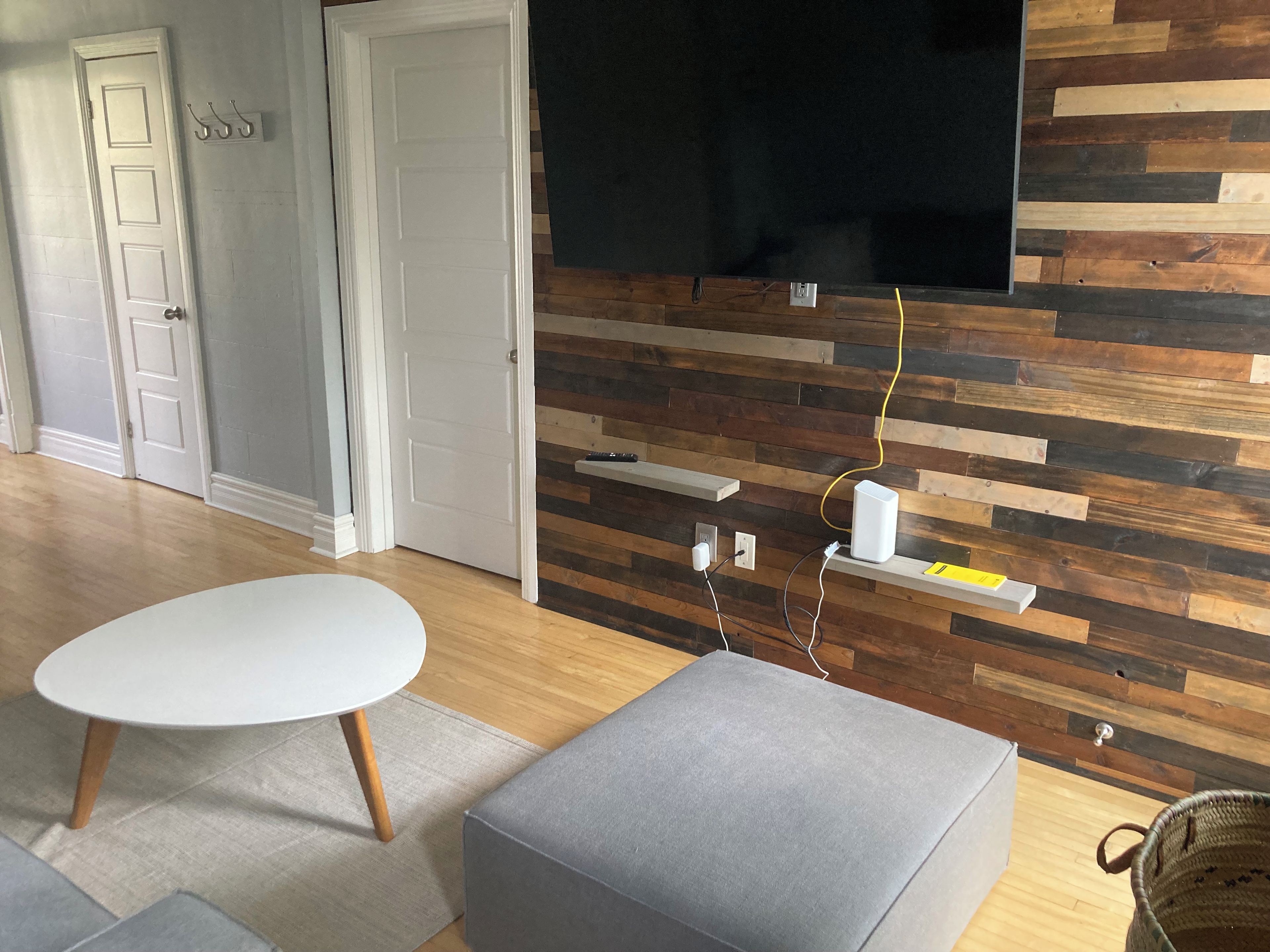 A modern living room with a flat-screen TV mounted on a wooden wall, a round white table, and a grey ottoman.
