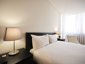 A hotel room with a bed, two bedside lamps, a nightstand with a remote control, and a chair. - FLH Boavista: Stylish Apartment with a View (Porto)