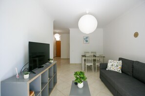 A modern living room with a flat-screen TV, a grey sofa, a dining table with chairs, and a potted plant. - FLH Olhão Minimalist Apartment with Balcony (Olhão)