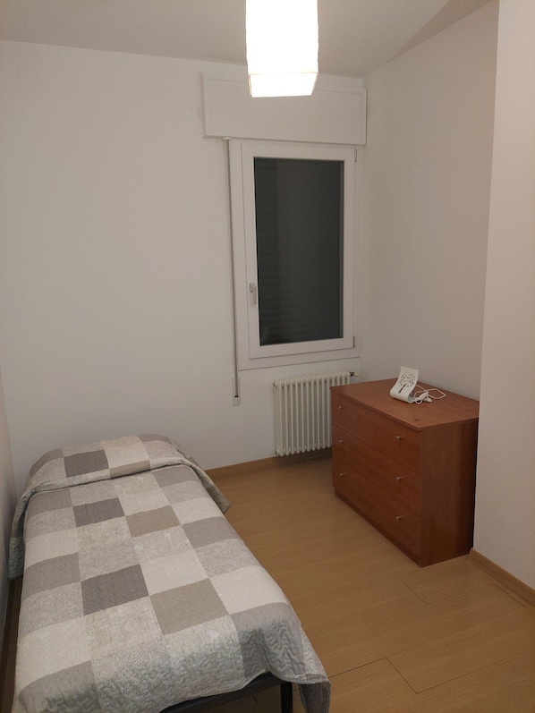 A small, sparsely furnished room with a single bed, a wooden dresser, a radiator, and a window.
