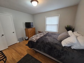 A bedroom with a bed, a dresser, a television, a window with blinds, and a door. - Cozy Allen Park Bungalow (Allen Park)