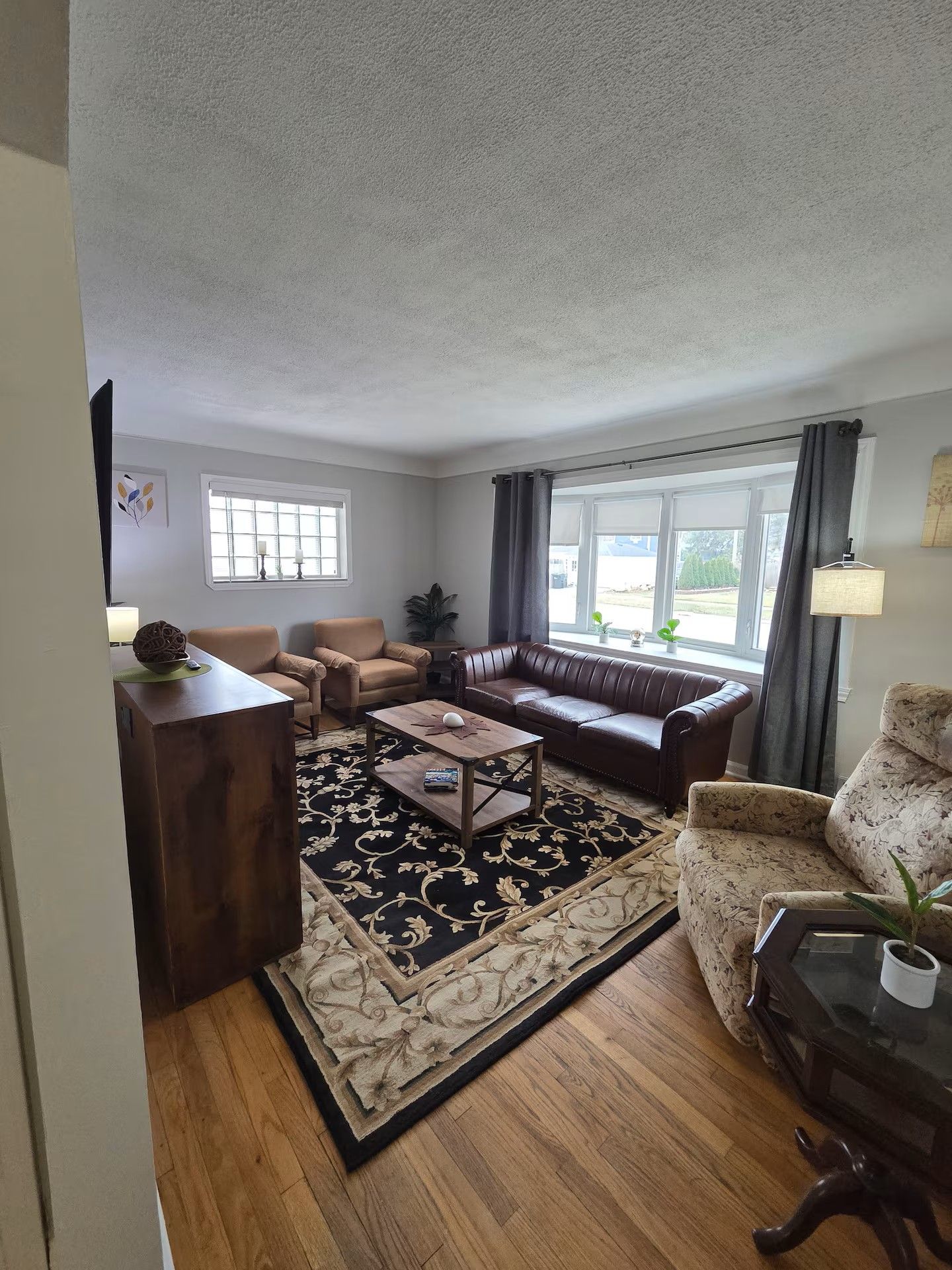Ein Wohnzimmer mit einem braunen Ledersofa, einem Holztisch und einem gemusterten Teppich. Zwei Fenster mit Vorhängen, ein Topf auf der Fensterbank und eine Lampe auf einem Beistelltisch.