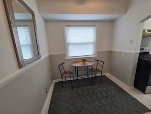 A compact kitchen with a dining table, two chairs, a window with blinds, and a refrigerator. - Cozy Allen Park Bungalow (Allen Park)